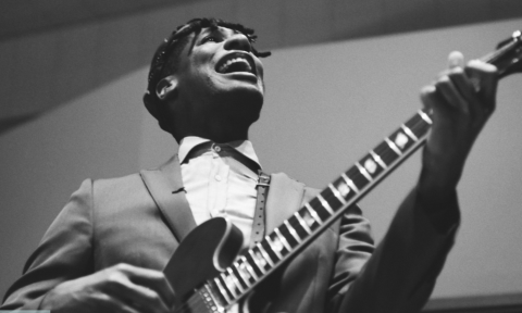 Jon Batiste smiles and stums his guitar.